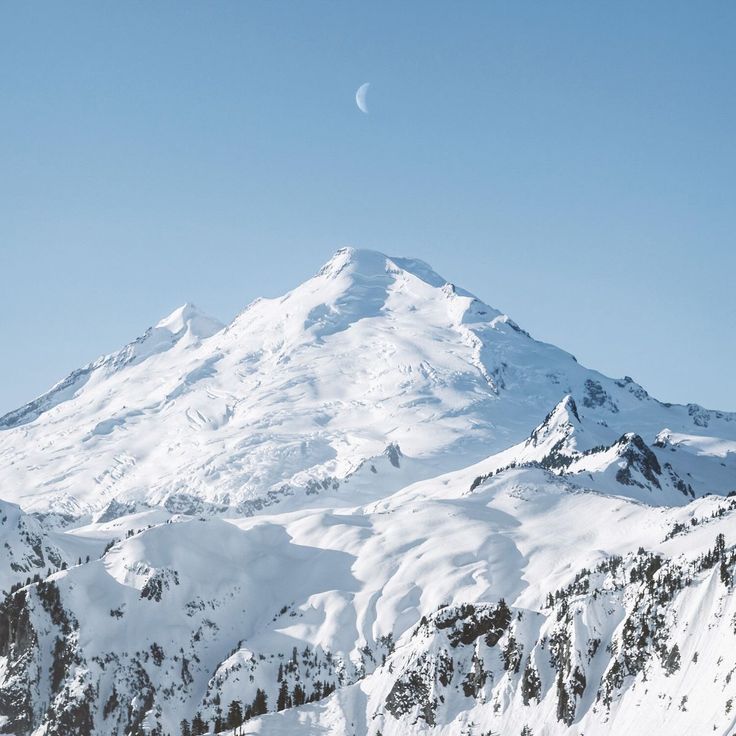 a mountain covered in snow under a blue sky with a half moon on it's side