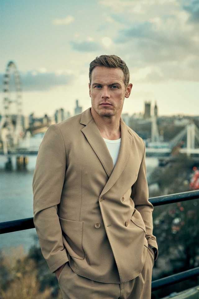 a man wearing a tan suit and white t - shirt standing in front of a cityscape