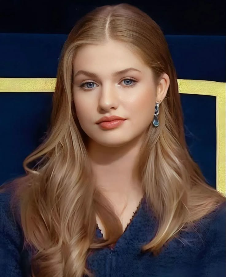 a woman with long blonde hair and blue dress sitting in a chair looking at the camera