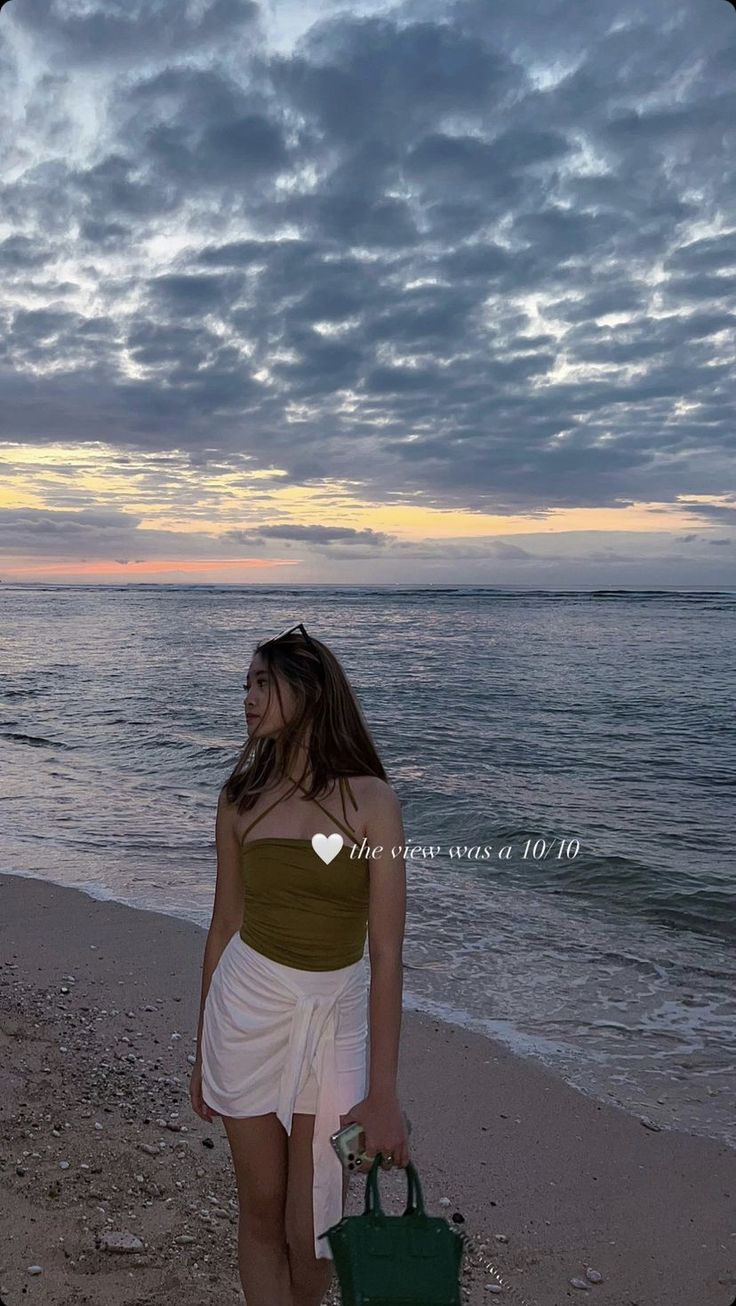 a woman standing on top of a beach next to the ocean holding a green purse