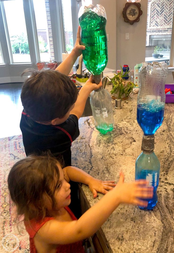 two children are sitting at a table with blue and green water in plastic bottles on it