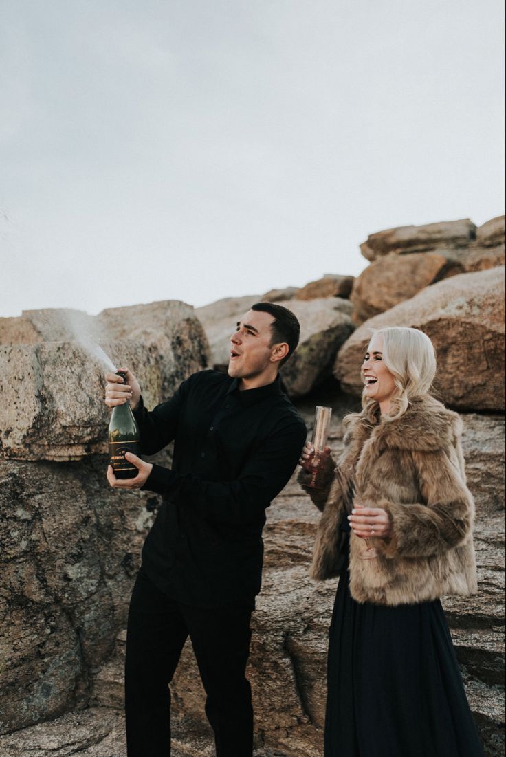 a man and woman standing next to each other near rocks with a bottle in their hand