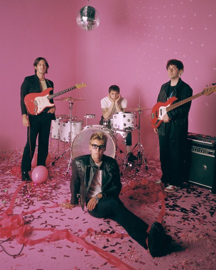 a group of people that are standing in front of some pink walls with guitars and drums