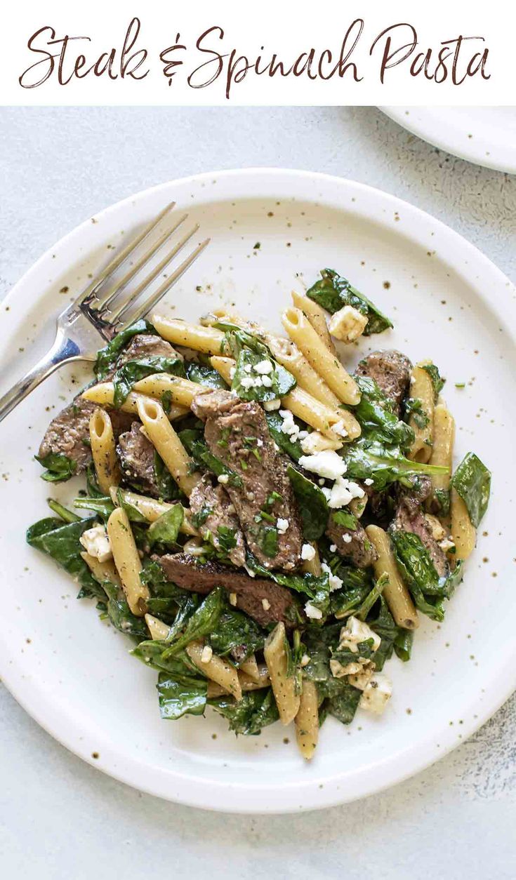 steak and spinach pasta on a white plate with a fork in the bowl next to it