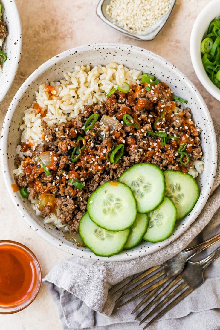 a bowl filled with rice and cucumbers next to bowls of salad, sauces and spoons