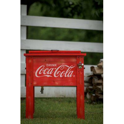 an old coca - cola cooler sitting in the grass next to a white fence and trees