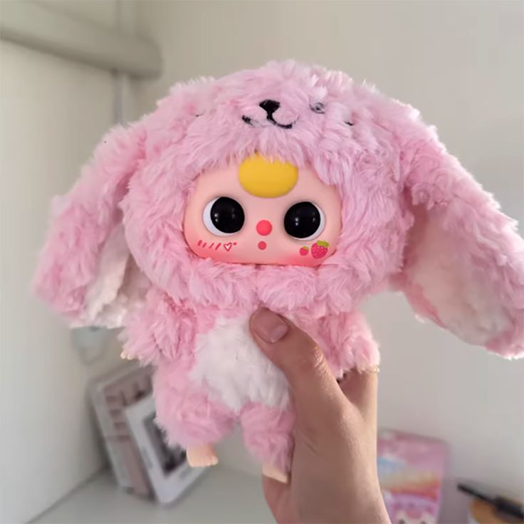 a hand holding a pink stuffed animal with black eyes and ears, in front of a white wall