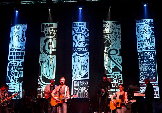 three men are playing guitars on stage in front of large windows with words projected behind them