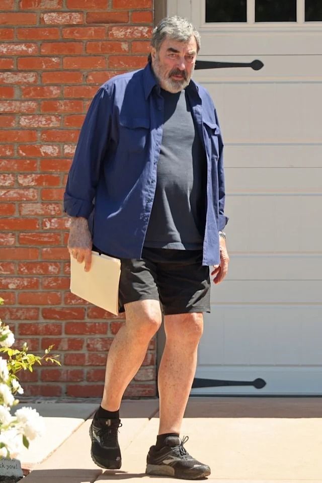 an older man in black shorts and a blue shirt is walking towards the camera with his hand on his hip