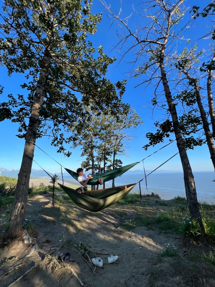 two people sitting in hammocks on top of a hill next to trees and the ocean