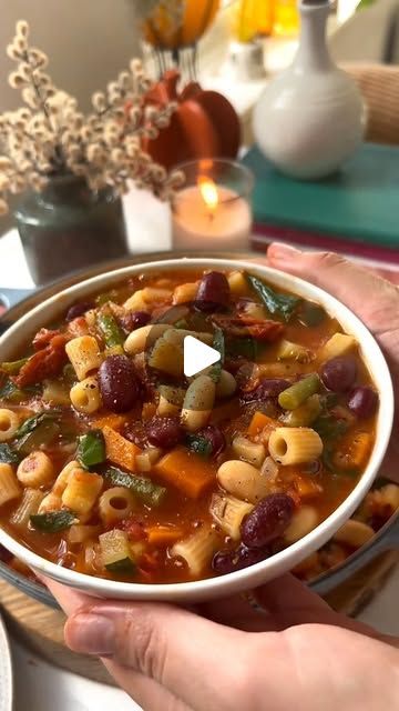 a person holding a bowl of soup with beans and veggies in it on a table