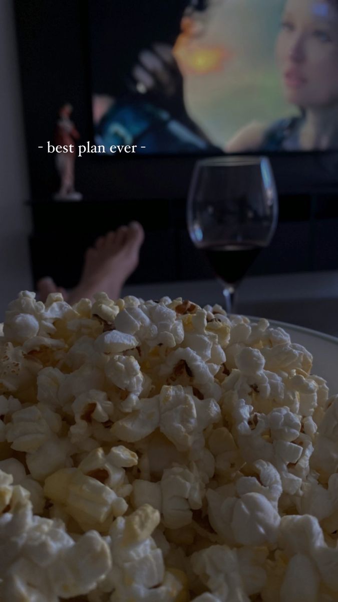 popcorn on a plate with a glass of wine in front of the tv and someone's feet