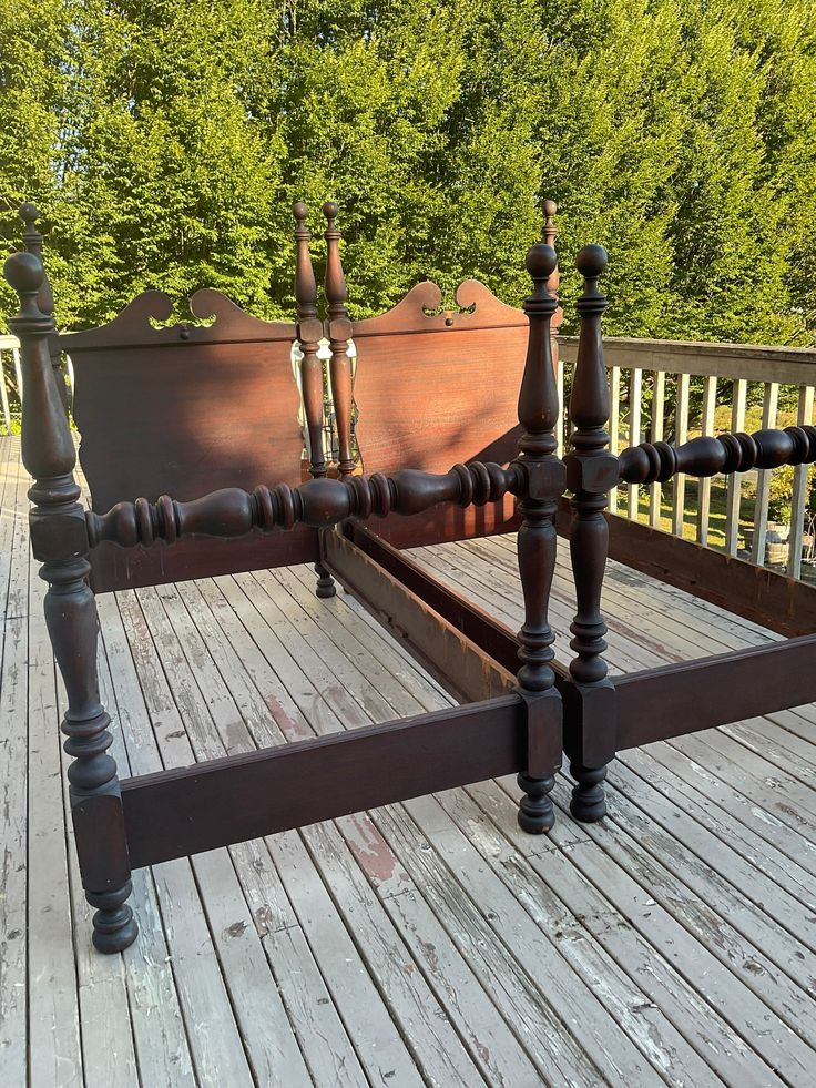 an old bed frame with wooden posts and turned down headboard on a deck in front of some trees