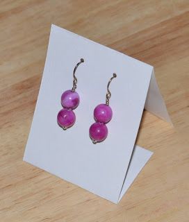 two pink beads are hanging from silver earwires on a white card next to a pair of scissors