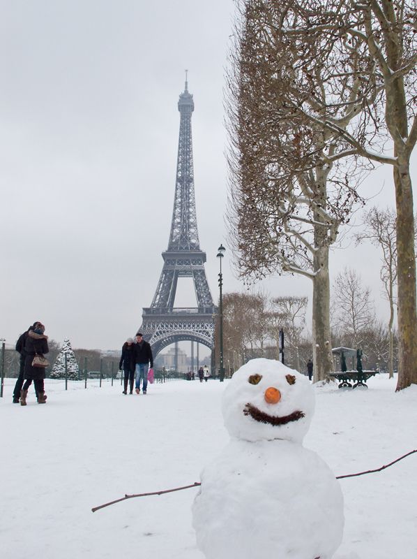 a snowman in front of the eiffel tower, with people walking by