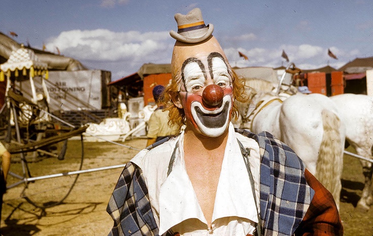 a clown with his face painted white and wearing a top hat, standing in front of some horses