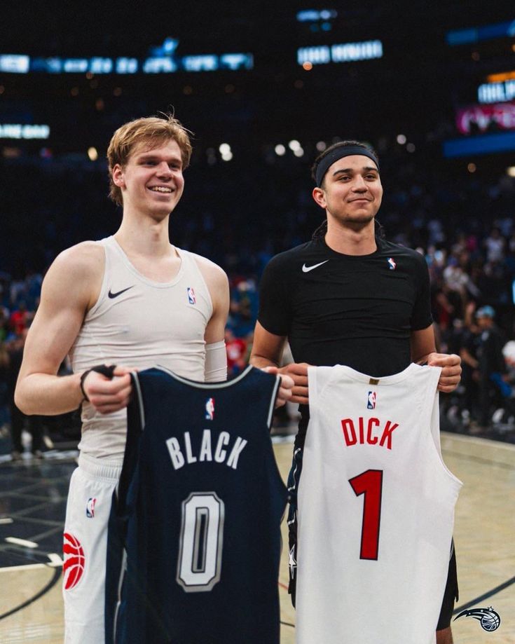 two young men holding up jerseys in front of a basketball court with fans watching from the stands