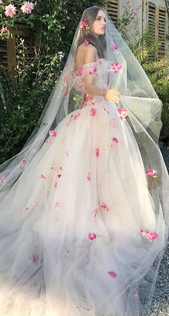 a woman in a wedding dress with pink flowers on the skirt and veil is standing outside