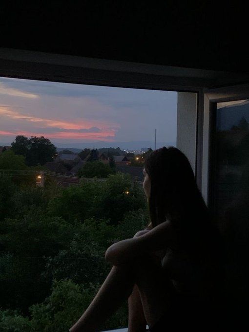 a woman sitting on a window sill looking out at the sunset