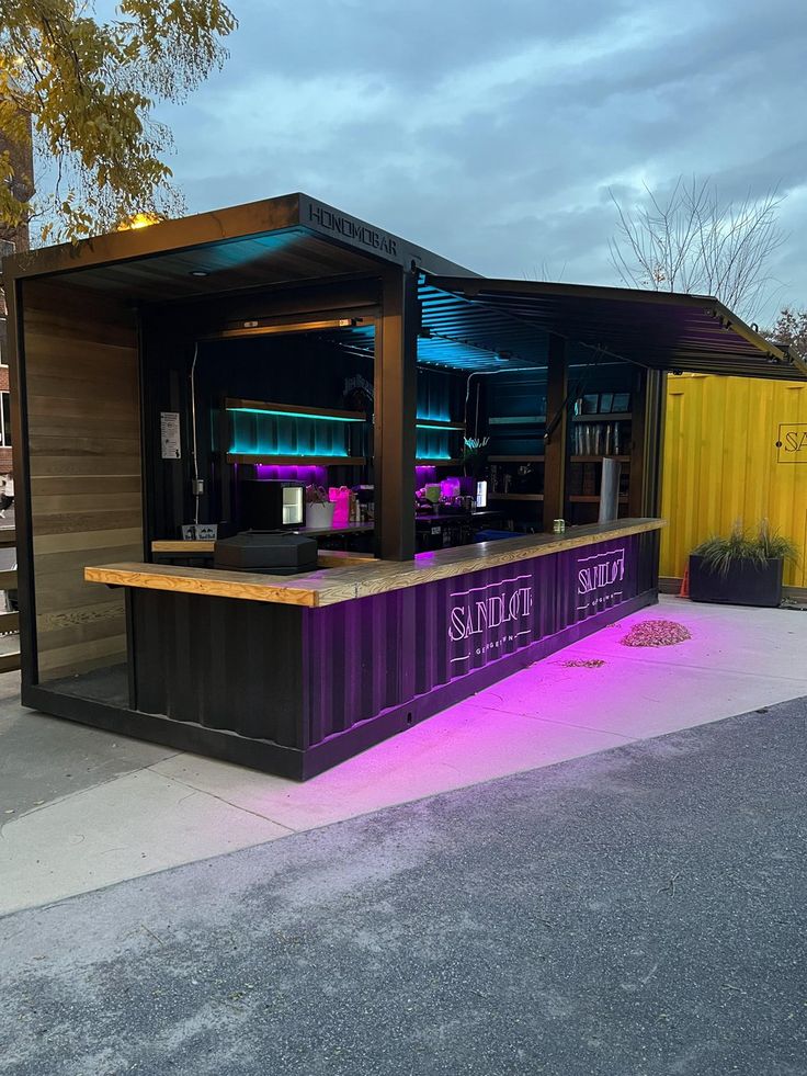 an outdoor bar is lit up with purple lights and sits next to a yellow fence