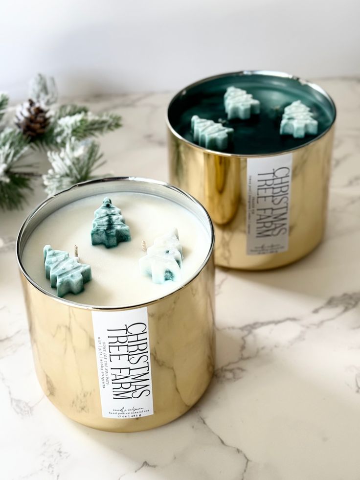 two tins filled with candles sitting on top of a marble counter next to evergreen branches