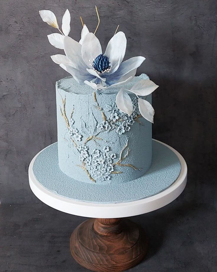 a blue and white cake with flowers on top is sitting on a pedestal in front of a gray wall