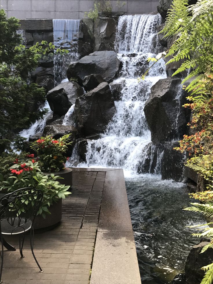 there is a waterfall in the middle of some plants and flowers next to a bench