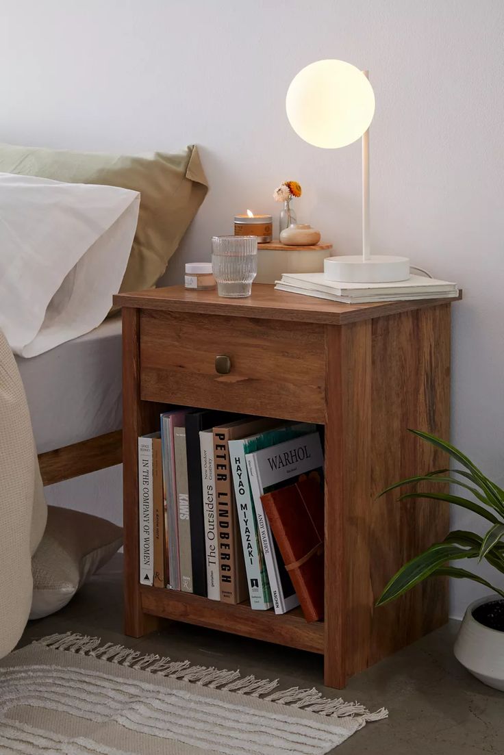 a nightstand with books on it next to a bed and a potted plant in the corner