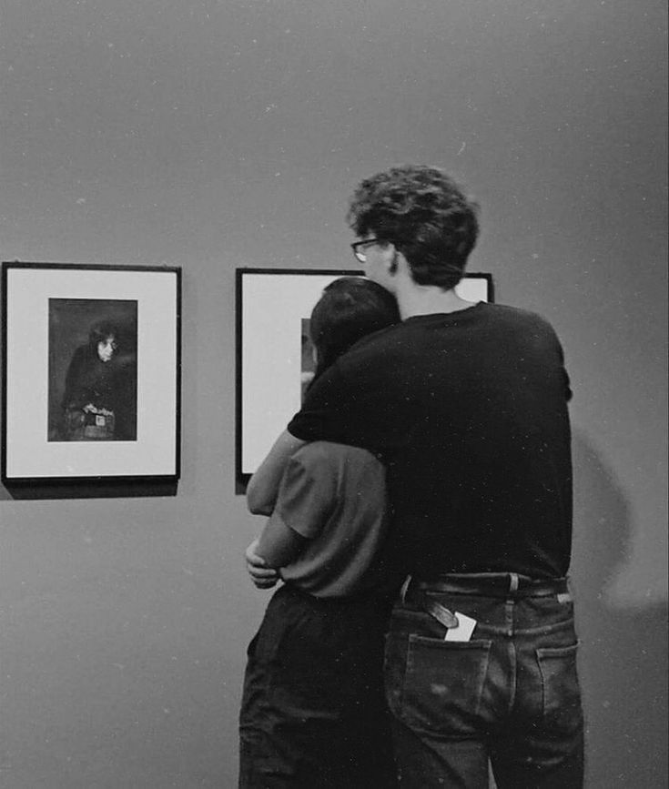 two people are looking at pictures on the wall in an art gallery, one is hugging the other's back