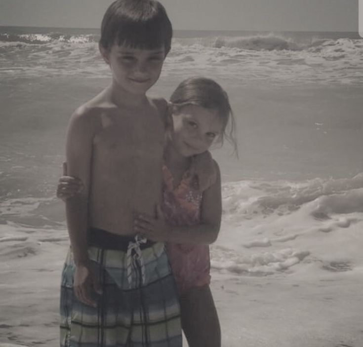 two young children standing on the beach in front of the ocean with their arms around each other