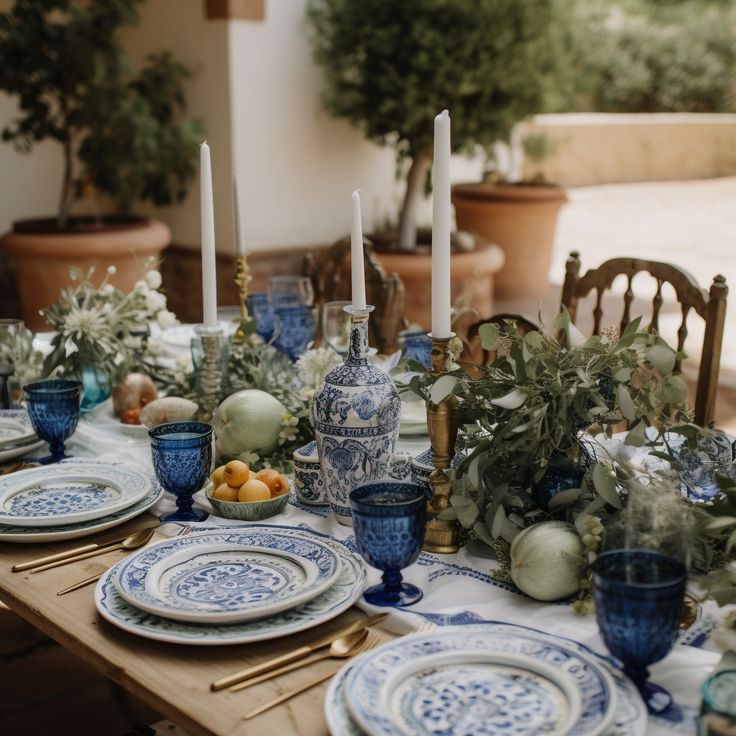 a table set with blue and white china plates, silverware, candles and greenery