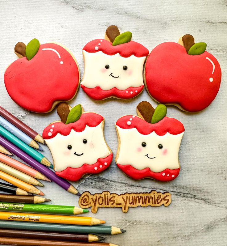 four decorated apple cookies sitting next to colored pencils