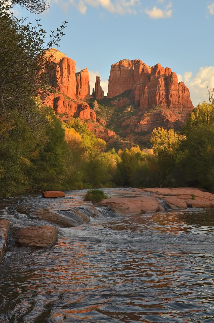 a river running through a lush green forest next to tall red rock formations in the background