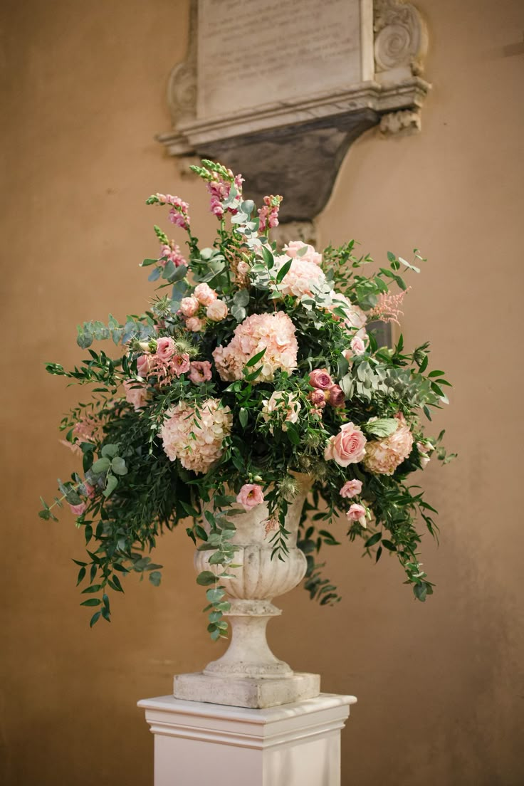 a vase filled with lots of pink flowers on top of a white pedestal next to a wall