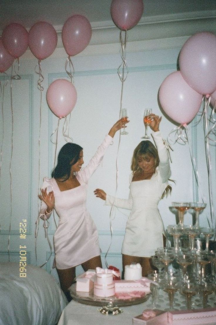 two women in white dresses are holding pink balloons and standing next to a table with cake on it