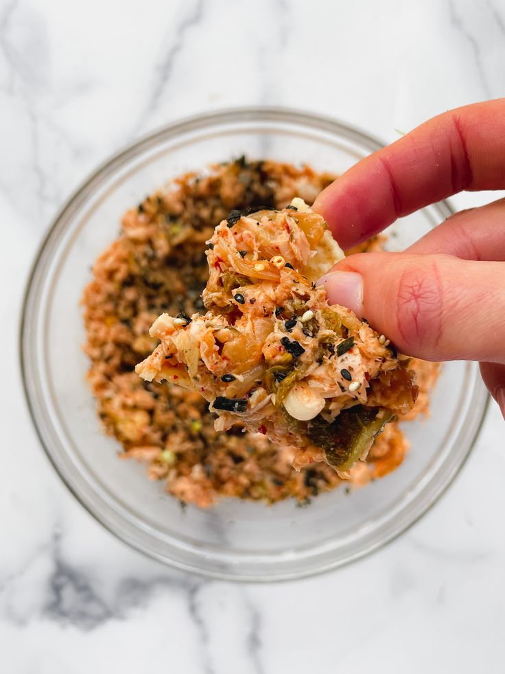 a hand holding a piece of food in a glass bowl on top of a marble counter