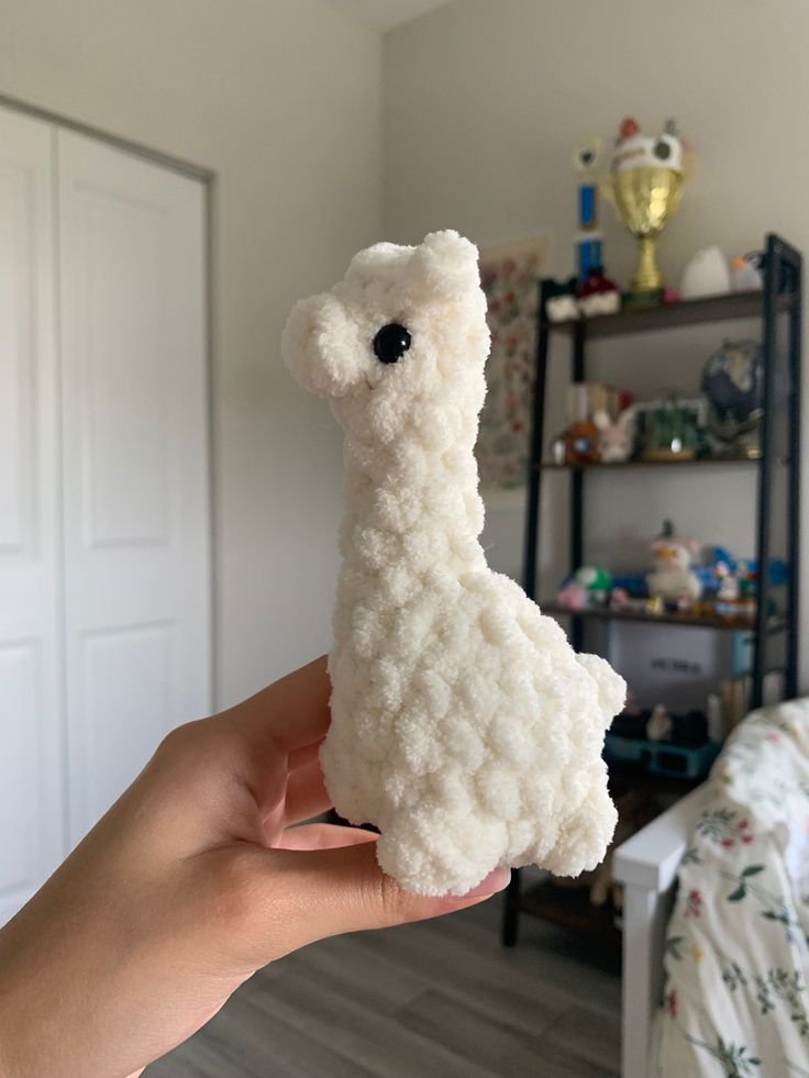 a hand holding a white stuffed animal in a room with a bed and shelving