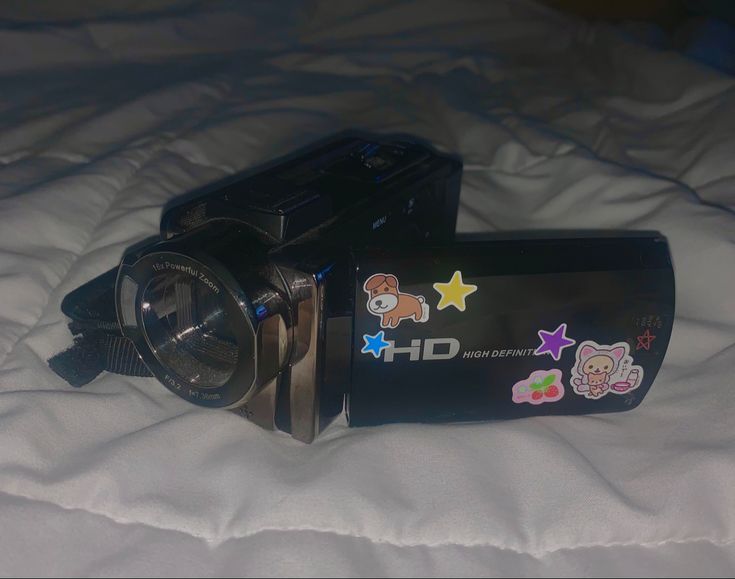 a black camera sitting on top of a white bed next to a teddy bear sticker