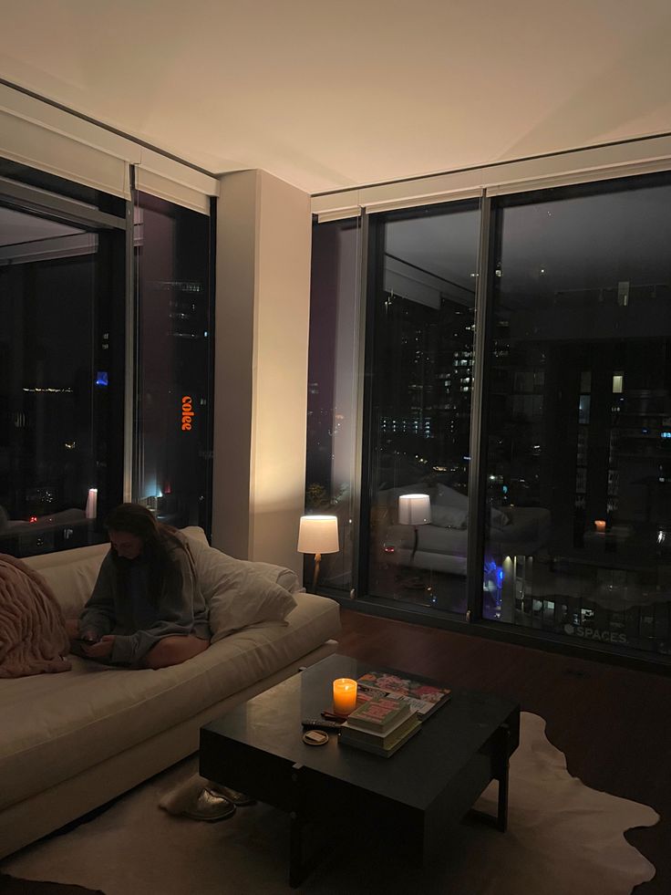 a woman sitting on a couch in front of a window with the city lights lit up