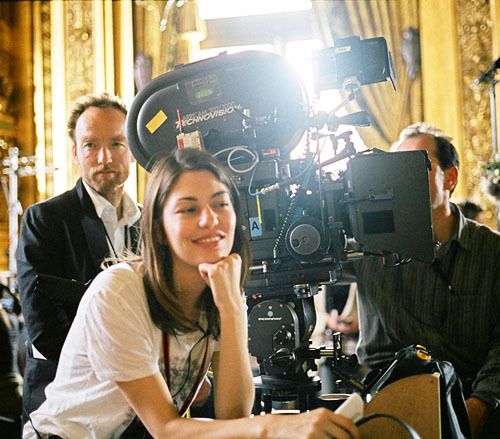 a woman sitting in front of a camera on top of a wooden table next to other people