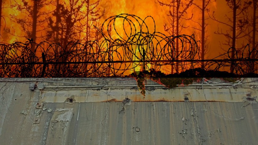 A razor-wire-topped prison wall with a wildfire raging in the sky behind it.
