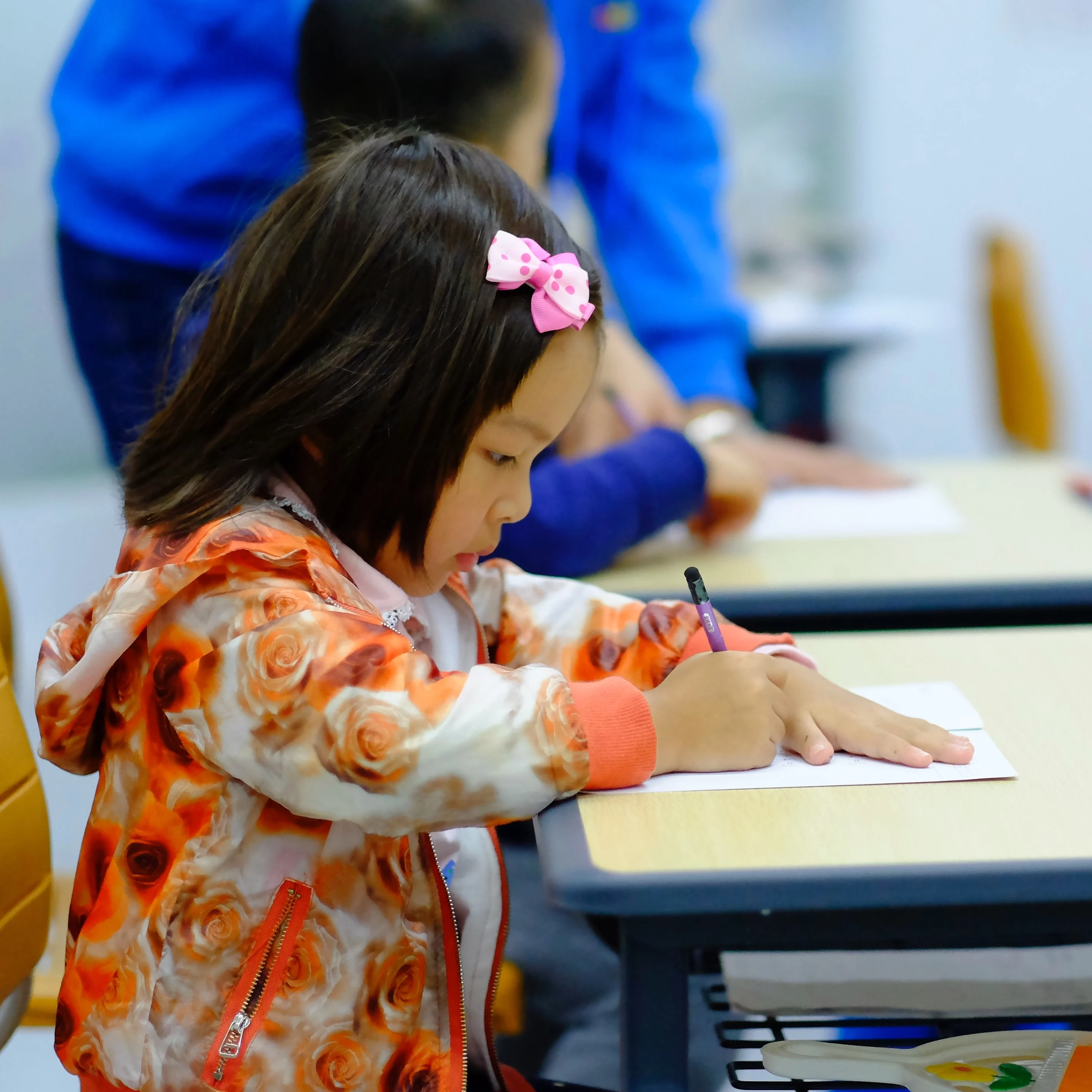 Young girl sits on a yellow school chair by a school table writing on a piece of paper