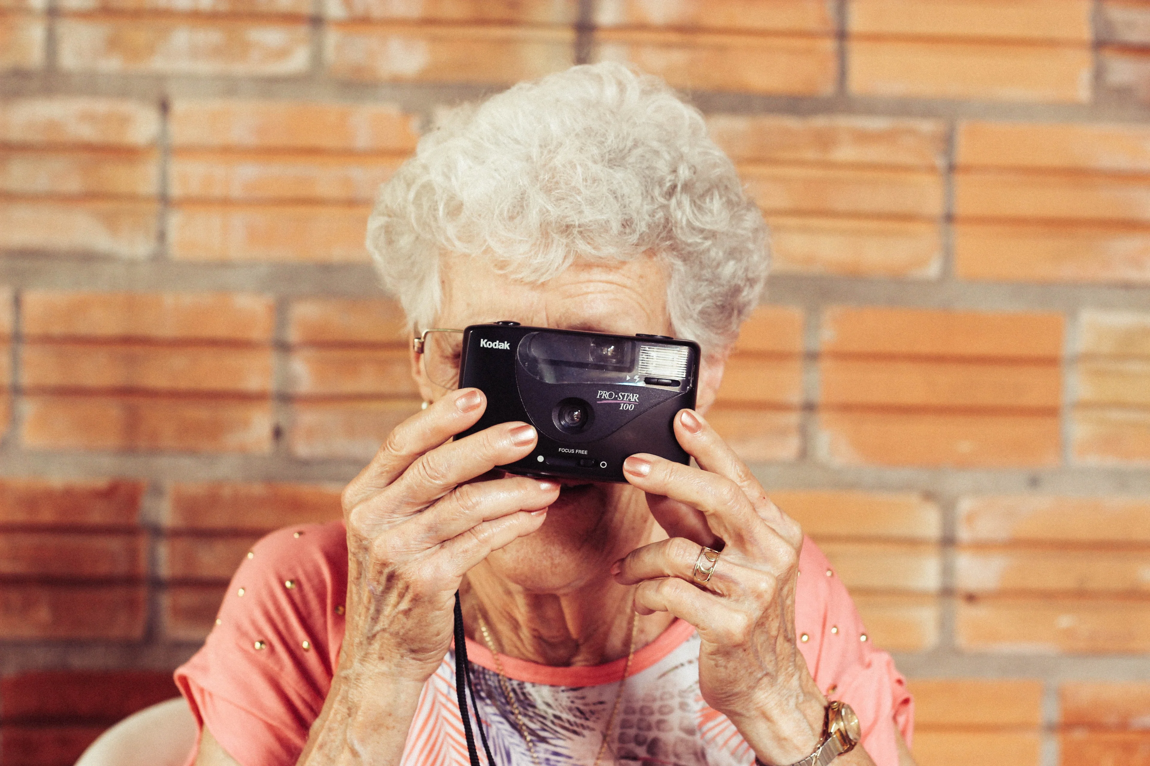 An older white haired woman in front of a brick wall gets ready to take a photo with a camera