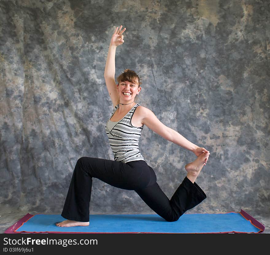 Young woman on yoga mat doing Yoga posture King Arthur's pose variation, with arm raised and hand in om position, looking at viewer and smiling, against a grey background in profile, facing left lit by diffused sunlight. Young woman on yoga mat doing Yoga posture King Arthur's pose variation, with arm raised and hand in om position, looking at viewer and smiling, against a grey background in profile, facing left lit by diffused sunlight.