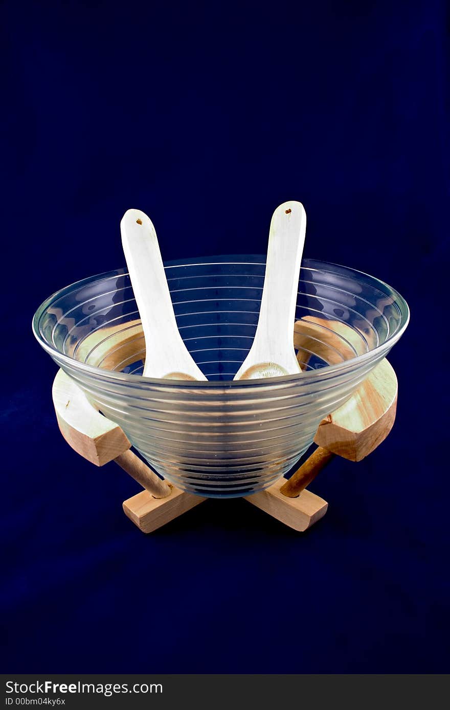 A glass salad platter with wooden cutlery and wooden support isolated on dark blue background.