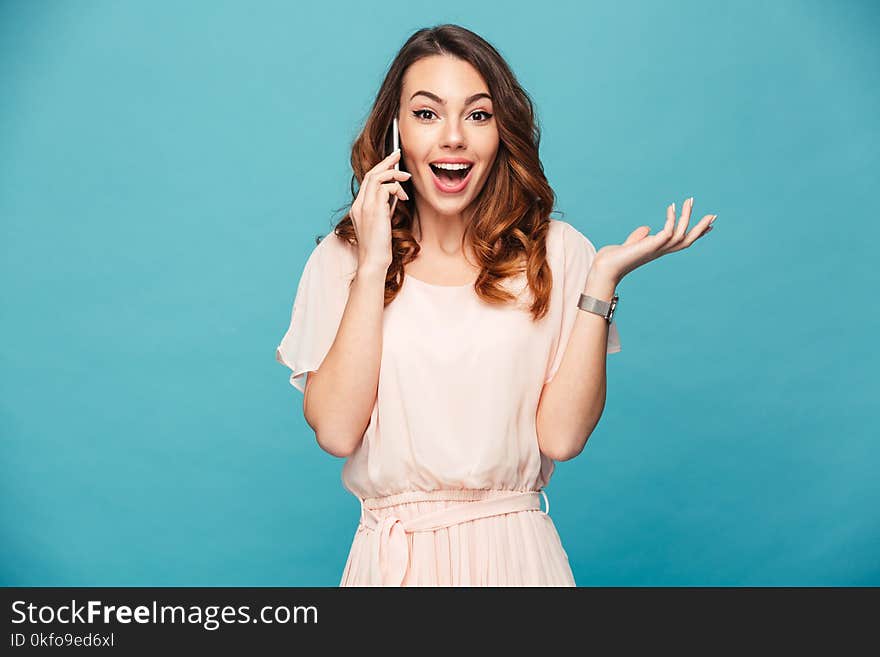 Portrait of a happy beautiful girl wearing dress talking on mobile phone and celebrating isolated over blue background