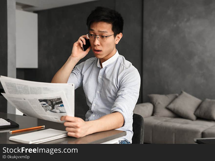 Portrait of an emotional young asian man talking by phone reading newspaper at home. Looking aside.