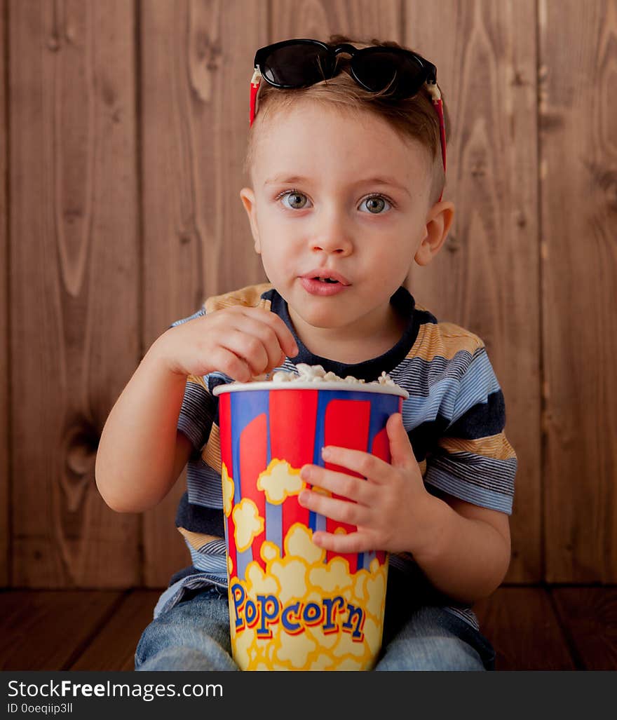 Little cute kid baby boy 2-3 years old , 3d cinema glasses holding bucket for popcorn, eating fast food on wooden background. Kids