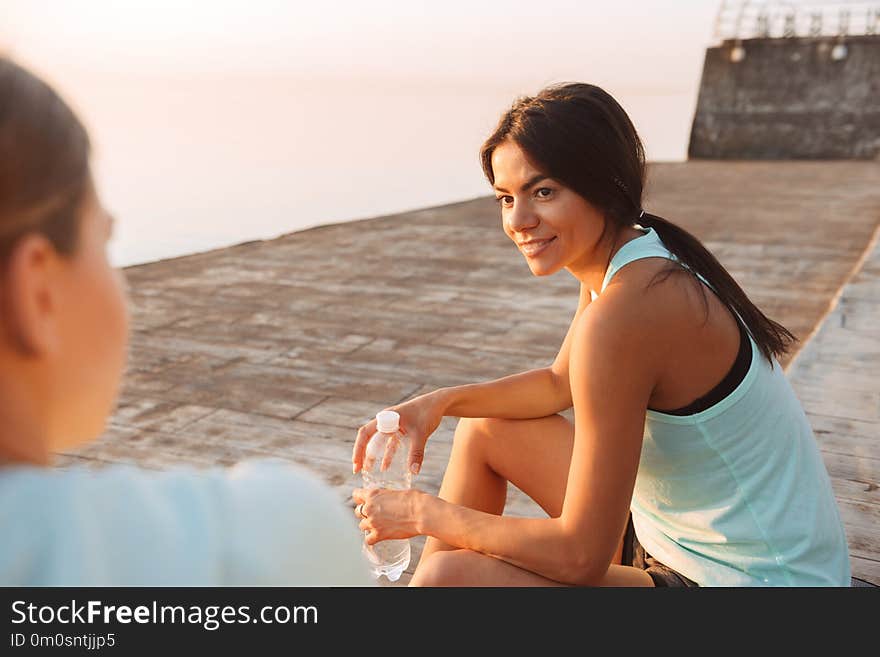 Picture of amazing young two sports women friends outdoors on the beach sitting talking with each other drinking water.