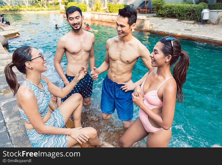 Multi-ethnic group of four young and cheerful friends smiling and talking while relaxing together at a trendy outdoor swimming pool in summer. Multi-ethnic group of four young and cheerful friends smiling and talking while relaxing together at a trendy outdoor swimming pool in summer